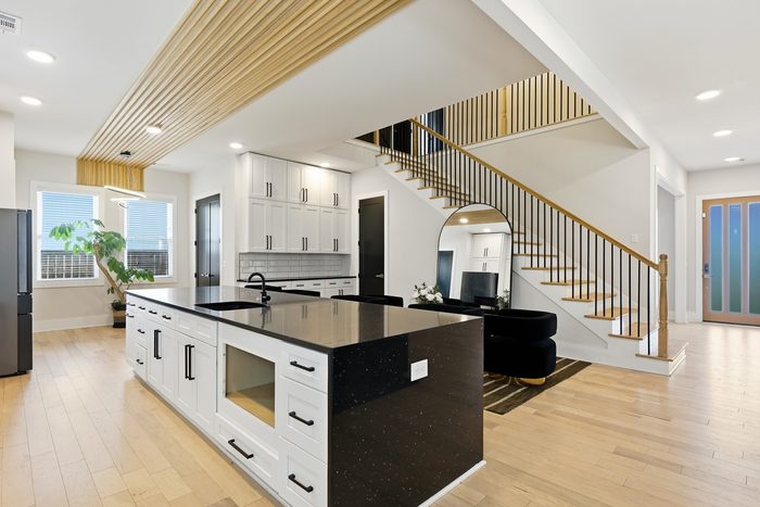 An open interior with a long black stone island, a slatted wood ceiling, an arched mirror and an open staircase with black balusters